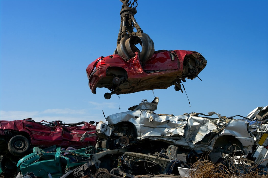 Red crushed car being picked up by a machine over pile of other wrecked cars.